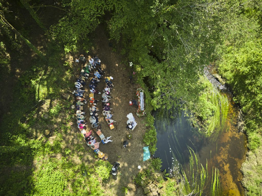 Vue aérienne drone d’un mariage en Bretagne avec invités réunis au bord d’une rivière entourée de forêt.