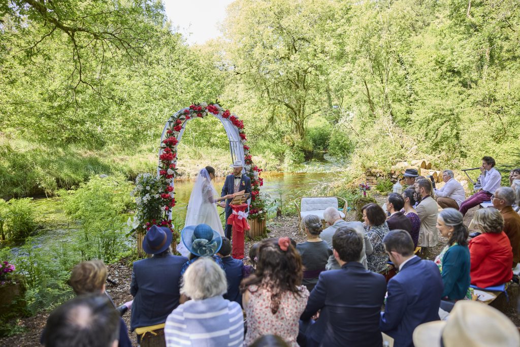 Mariés échangeant leurs vœux sous une arche fleurie au bord d’une rivière lors de leur mariage en Bretagne.